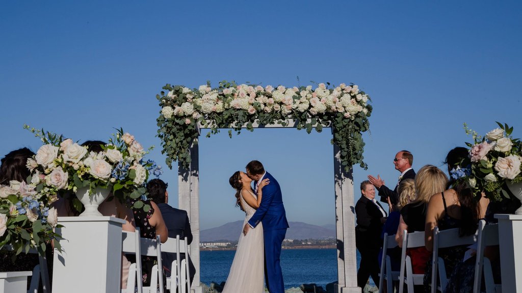 Bride and groom kissing under a white floral arch at a seaside ceremony as guests applaud, showing how relationship quality helps predict infidelity