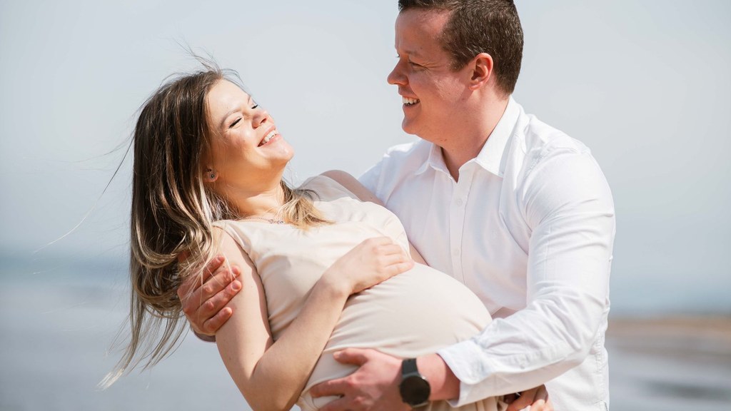 Smiling man embracing his pregnant partner on a bright beach, showing acceptance of body change, body image and sexual satisfaction