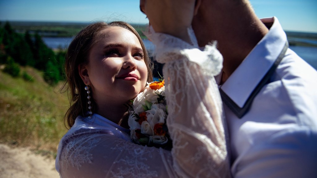 Bride smiling up at her groom in a close outdoor embrace, kissing and partner choice in a lasting marriage