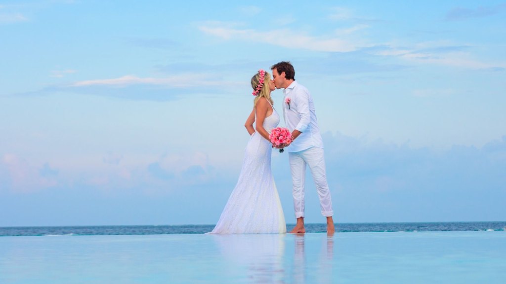 A bride with a pink flower crown and a clean-shaven groom in white kiss by the sea under a blue sky, newlyweds who value sexual communication