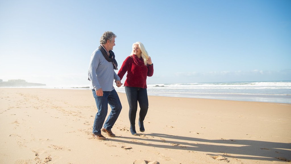 Couple in their fifties holding hands and walking along a sunny beach, talking about changes in sexual desire at midlife and low sex drive in your 50s