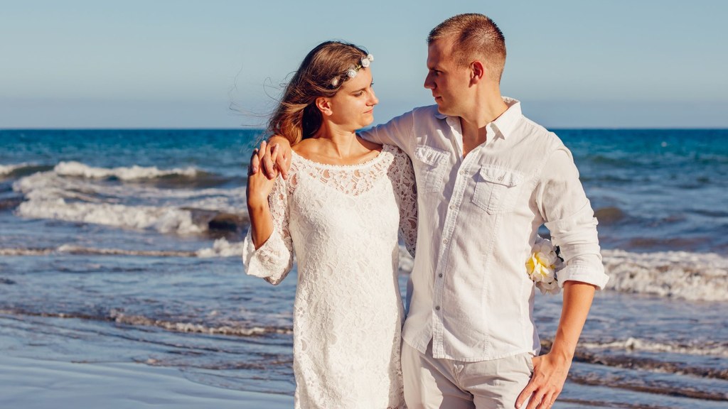 Newlywed couple in white linen standing arm in arm on a sunny beach, illustrating how often married couples have sex as the years pass