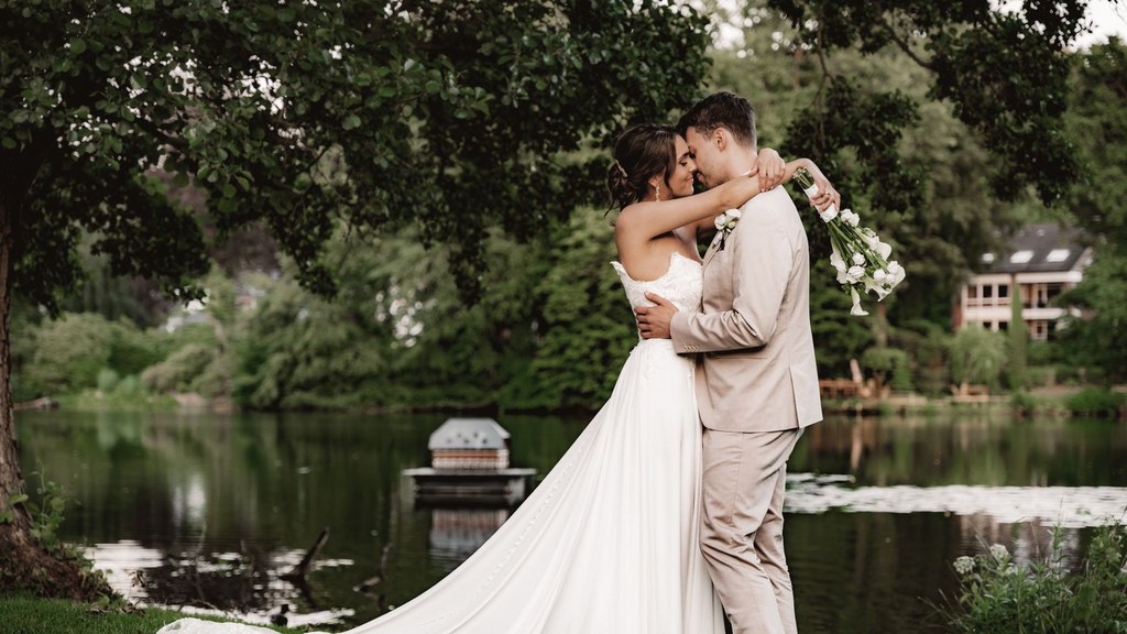 Bride and groom embracing by a green lakeside on their wedding day, hugging and health in a committed bond