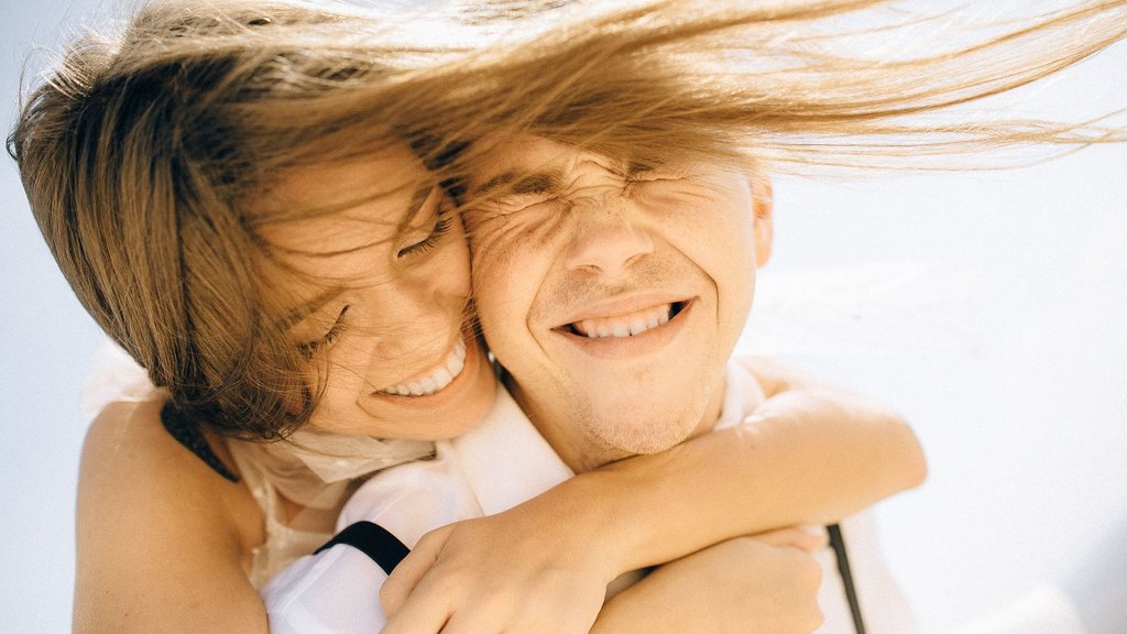 A laughing couple in a tight, cheek-to-cheek embrace under bright sun, hair blowing in the wind, illustrating couple similarity and partner choice