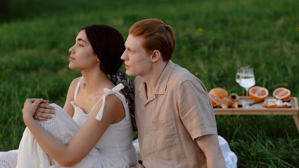 Mental fatigue and relationships: a couple sitting close on summer grass while she looks away, lost in thought after a stressful day
