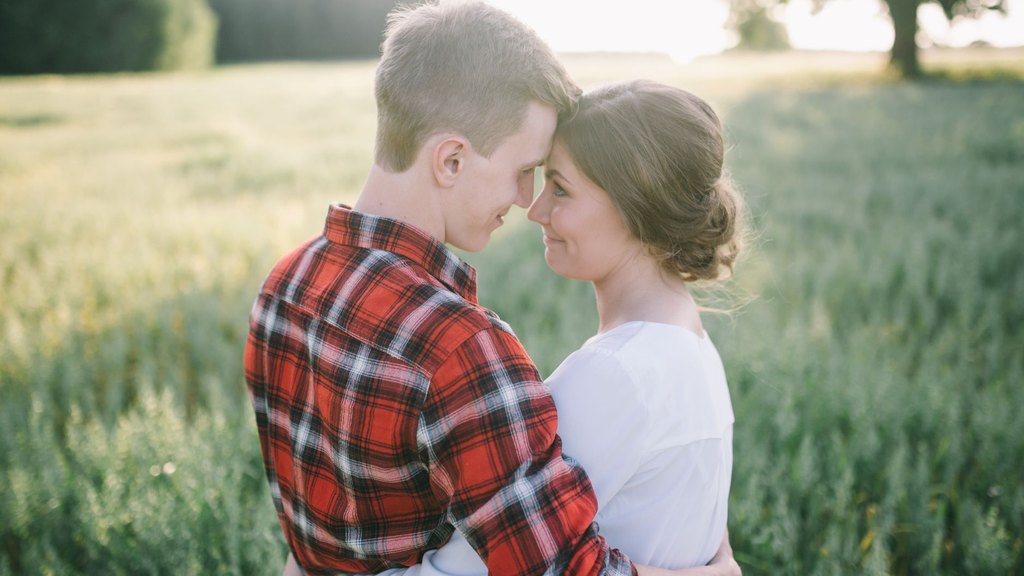 A clean-shaven young man in a plaid shirt and a woman in a white blouse touch foreheads in a sunny green field, illustrating couple similarity on a first date